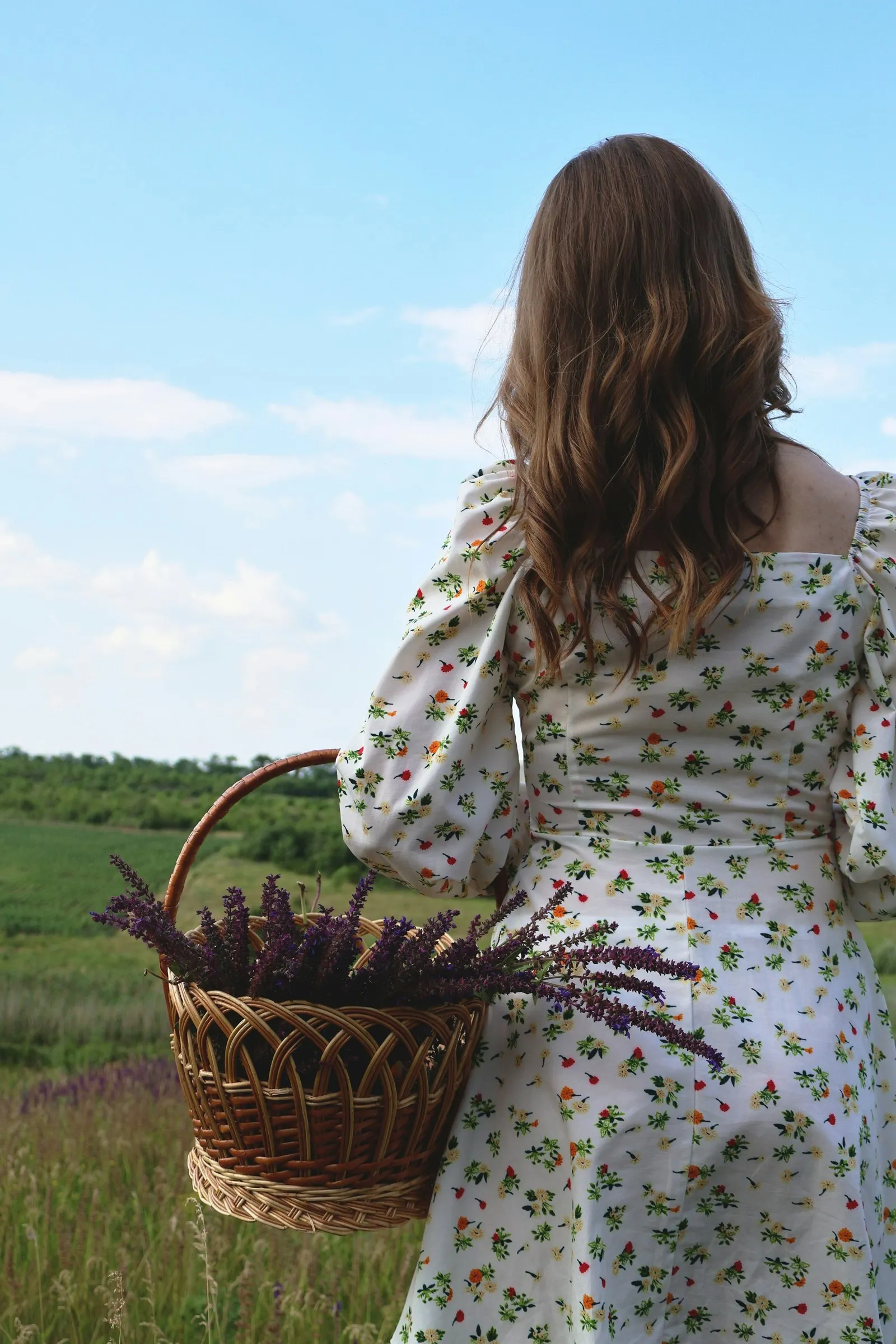 Mulher de costas em um campo, com vestido floral de mangas compridas e uma cesta de lavanda