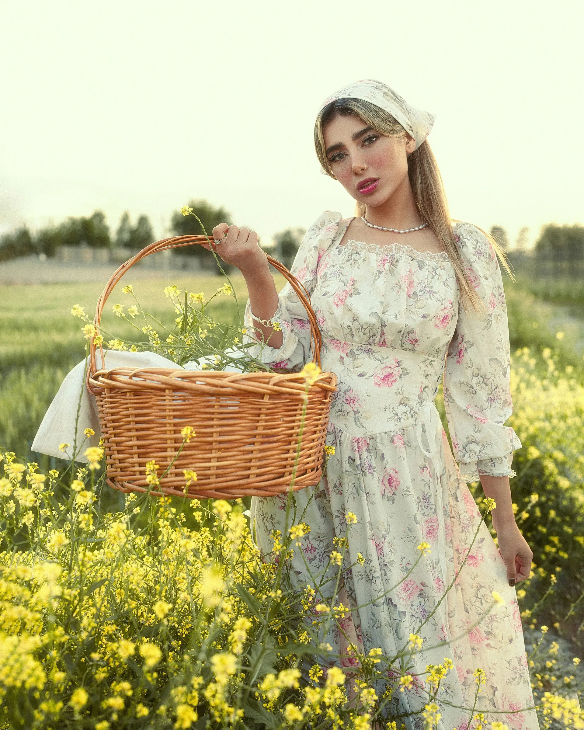 Mulher sorrindo em um campo de flores amarelas, com lenço no cabelo, vestido longo floral de mangas compridas e uma cesta de flores