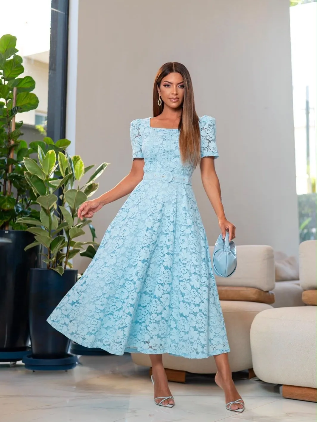 Vestido Silvana azul-claro em renda, com decote quadrado e cinto, em um ambiente interno claro